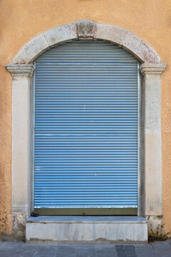 Weathered arched doorway featuring a closed light blue metal roller shutter, framed by aged white stone columns and a textured ochre plaster wall on an old building exterior.