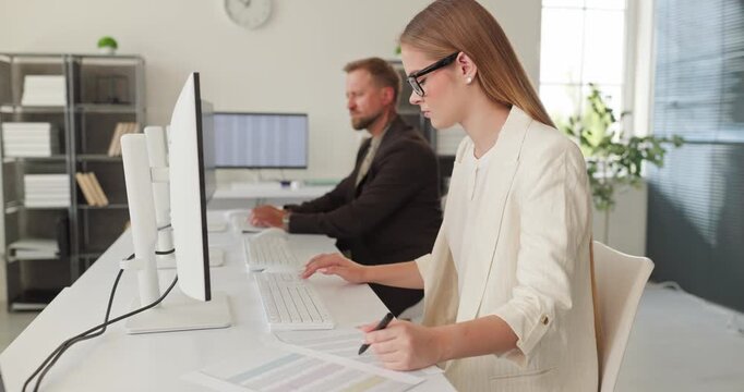Accountant woman at computer in office. Focused analysis of database and report on dual monitors with colleagues nearby and teamwork enabled by technology. Theme of finance and productivity.