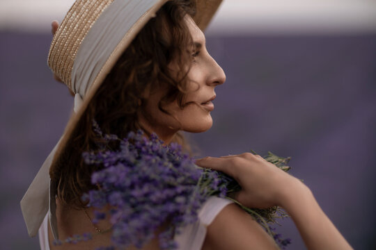 Straw hat wearing young woman holds lavender bouquet in rural field during summer sunset