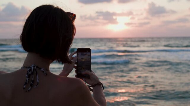 The beach, phone and woman take photos of the sunset for social media during an evening walk on the beach of Israel. The girl is holding her phone and filming the content. sunset at the beach