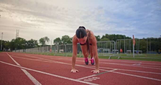 Young athletic woman in sportswear warming up and doing exercises on the stadium running track in the evening at sunset