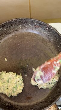 Zucchini fritters cooking in a pan. Overhead high-angle close-up shot of a wooden spatula pressing raw batter into a hot metal skillet.