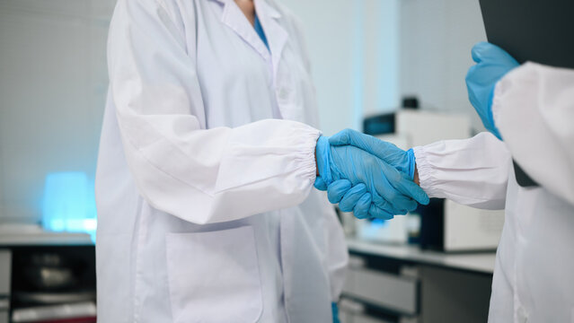 Close up of Two Scientists Shaking Hands Wearing Blue Latex Gloves and White Lab Coats in Modern Laboratory