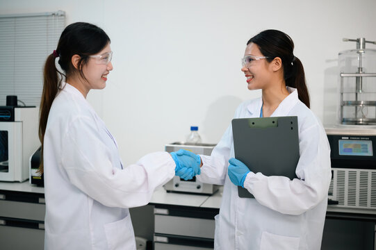 Two Female Scientists Shaking Hands in Laboratory, Wearing White Lab Coats and Safety Goggles