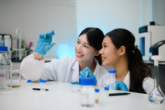 Two Young Asian Female Scientists Smiling and Examining Sample Vial Together in Modern Laboratory