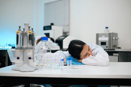 Exhausted Female Scientist Sleeping at Laboratory Bench Surrounded by Test Tubes and Equipment