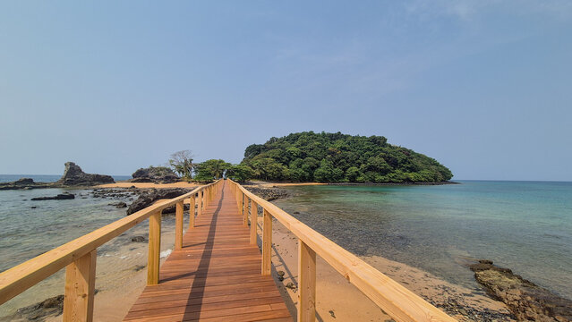Wooden boardwalk leading to Bom Bom Islet on Pr&iacute;ncipe Island, S&atilde;o Tom&eacute;. Scenic coastal view featuring a pedestrian bridge above crystal-clear waters, connecting the tropical shoreline to the islet