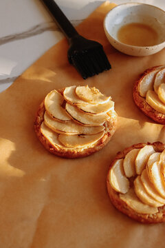 Close-up of freshly baked mini pear and frangipane tart brushed with glaze using pastry brush, small bowl of glaze on baking paper in warm evening light