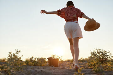 Fototapeta premium Young woman in a striped top and white shorts walks away at sunset, holding a wide hat, strolling along a dusty path as warm light glows on her silhouette.