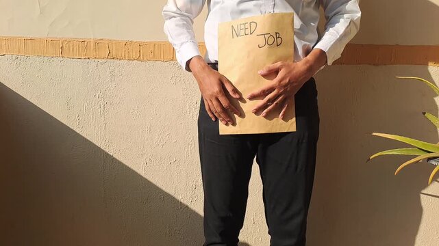 A job seeker wearing a white shirt and black pants holding a brown envelope with a handwritten need job text. Unemployment, career, and recruitment stock video footage.