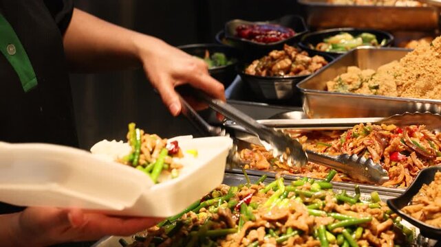 Hands use tongs to fill a takeout container with stir fried food