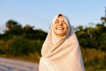 Fototapeta premium Smiling woman wrapped in a light shawl outdoors on a sunny day, enjoying a gentle breeze. Lifestyle portrait for wellness, travel, or nature themes.