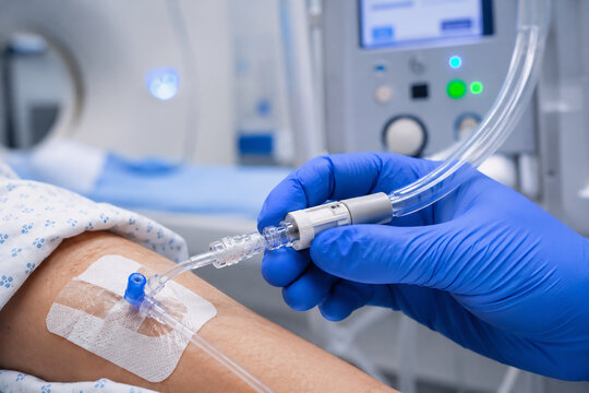 Close-up shot of an imaging technician's gloved hand connecting an automatic power injector tube to a patient's IV access line for contrast-enhanced CT scan, modern radiology room background