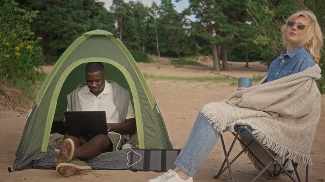 Young adult African American man working on laptop inside camping tent while young adult Caucasian woman sits nearby on beach. Perfect for remote work and digital nomad lifestyle commercials