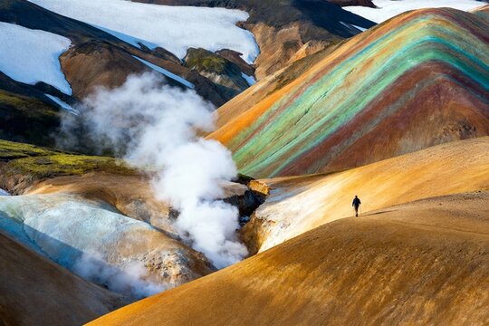 A lone hiker traverses a vibrant, geothermally active mountain landscape with colorful sediment layers and steam vents.