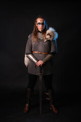 Fototapeta premium Young Viking woman with combat makeup on her face in chainmail with fur and a sword in her hands posing while standing against a dark background with blue backlighting.