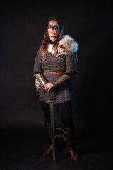 Fototapeta premium Young Viking woman with combat makeup on her face in chainmail with fur and a sword in her hands posing while standing against a dark background with blue backlighting.