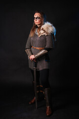 Fototapeta premium Young Viking woman with combat makeup on her face in chainmail with fur and a sword in her hands posing while standing against a dark background with blue backlighting.