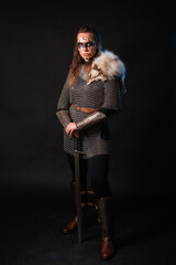Fototapeta premium Young Viking woman with combat makeup on her face in chainmail with fur and a sword in her hands posing while standing against a dark background with blue backlighting.