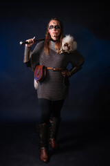 Fototapeta premium Young Viking woman with combat makeup on her face in chainmail with fur and a sword in her hands posing while standing against a dark background with blue backlighting.