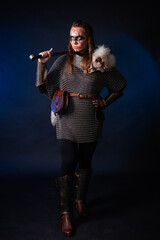 Fototapeta premium Young Viking woman with combat makeup on her face in chainmail with fur and a sword in her hands posing while standing against a dark background with blue backlighting.