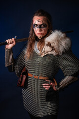 Fototapeta premium Young Viking woman with combat makeup on her face in chainmail with fur and a sword in her hands posing while standing against a dark background with blue backlighting.