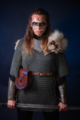 Fototapeta premium Young Viking woman with combat makeup on her face in chainmail with fur and a sword in her hands posing while standing against a dark background with blue backlighting.