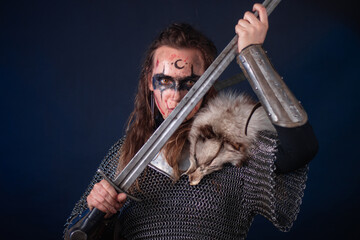 Fototapeta premium Young Viking woman with combat makeup on her face in chainmail with fur and a sword in her hands posing while standing against a dark background with blue backlighting.
