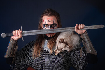 Fototapeta premium Young Viking woman with combat makeup on her face in chainmail with fur and a sword in her hands posing while standing against a dark background with blue backlighting.