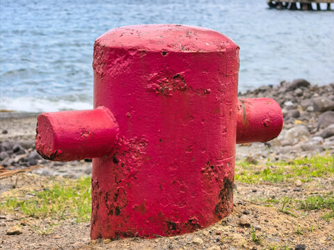 Weathered red metal mooring bollard installed on a coastal shore by the sea.
