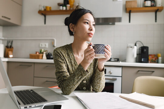 Pensive dreamy asian girl having coffee break during her freelance remote work as accountant, holding mug of hot drink in hands and looking aside at window, relaxing and contemplating at kitchen table
