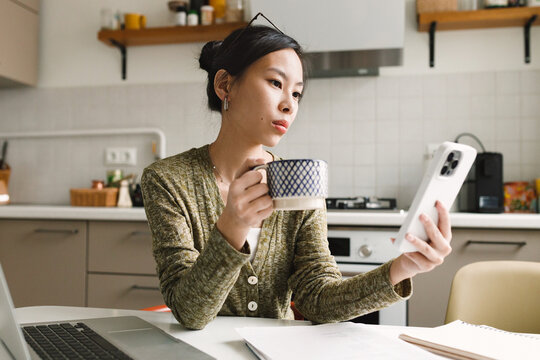 Young asian woman having online conversation with ai chat bot via mobile application, reading for its detailed answer for her personal question, while drinking coffee sitting at kitchen table