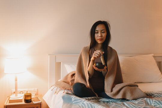 Calm meditating young asian girl with blanket on shoulder holding singing copper bowl and stick to get healing sound sitting on bed in evening with closed eyes, enjoying spiritual practice at home