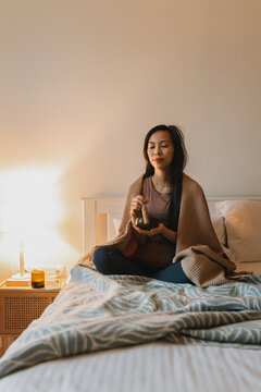 Vertical image of asian woman with singing bowl on bed with eyes closed, wrapped in blanket. Healing sound vibrations promoting relaxation, mental clarity and spiritual wellness at home before sleep