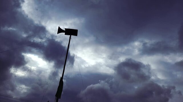 Wide silhouette of a loudspeaker horn with a flag fluttering on a pole under deep blue storm clouds, capturing a sense of urgent community broadcast, calling to prayer, or impending harsh weather.