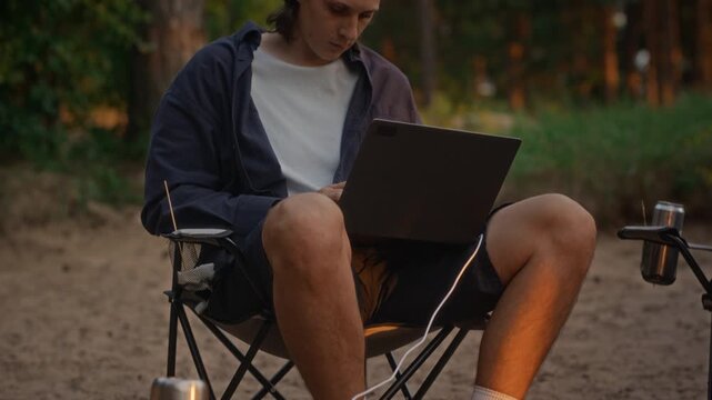 Caucasian young adult man working on laptop while camping on beach, camera tilting up from campfire. Footage ideal for remote work, digital nomad lifestyle, and off-grid technology commercials