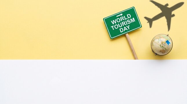 World Tourism Day concept with green sign, globe, and airplane shadow. Minimalist flat lay on yellow background for travel promotion.