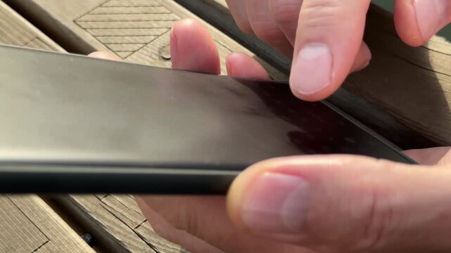 Male hands typing message on smartphone outdoor while sitting at wooden table. Fingers holding black mobile device sending quick digital text in chat app. Outdoor communication concept with
