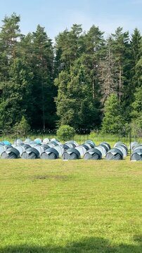 Rows of grey camping tents setup on green grass field near woodland. Tourist rally base camp located on forest meadow during warm summer day. Outdoor travel expedition and group camping trip