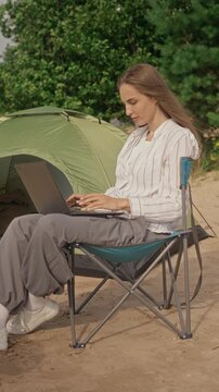 Vertical shot of young adult Caucasian woman sitting in folding chair near camping tent on beach, typing on laptop. Clip is ideal for promoting digital nomad lifestyle and remote work freedom