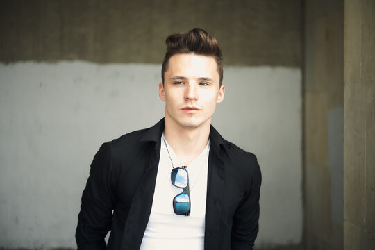 Close-up portrait, man in black shirt over white tee, sunglasses on neck, looking aside. Serious expression, styled hair, confident editorial fashion look. Two-tone wall background.