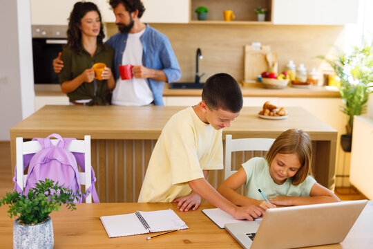 Two siblings working together on schoolwork with a laptop while their parents spend time in the kitchen behind them