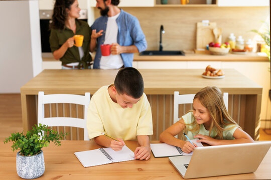 Two children doing homework at a kitchen table while their parents talk in the background