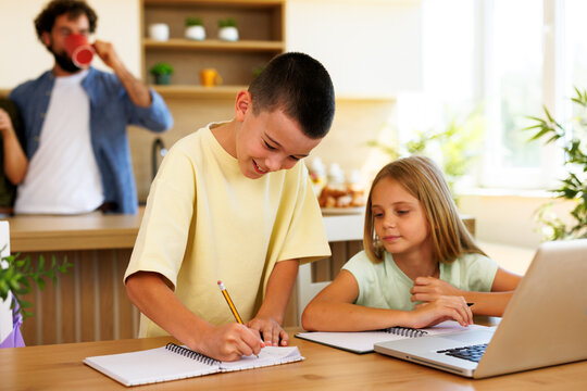 Young boy writes in a notebook while his sister works beside him with a laptop during homework time