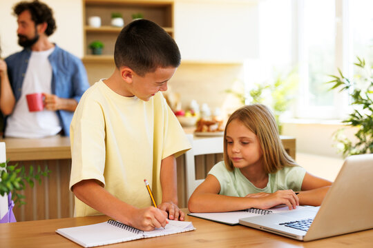 Two siblings work together on their homework at a kitchen table while a parent relaxes in the background