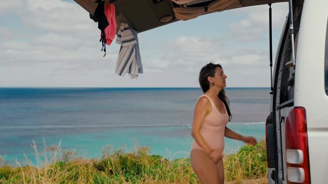 Woman in a swimsuit removes her surfboard from the back of a parked white van during a sunny afternoon trip to the coast