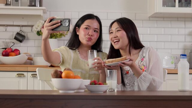 Two women take a selfie together while enjoying food and drinks in a bright modern kitchen. Ideal for friendship, social media, lifestyle, home, food, and digital communication concepts.
