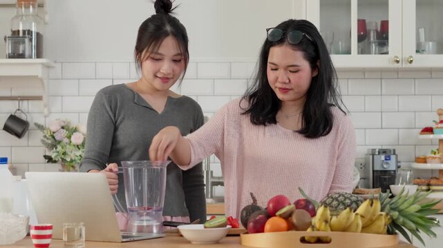 Asian women prepare a fresh fruit smoothie together in modern kitchen, blending healthy ingredients while enjoying a relaxed lifestyle moment. Ideal for wellness, nutrition and healthy living concepts
