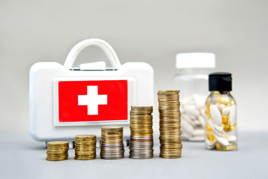 Stacks of coins rising in front of a first aid kit and bottles of medication. Rising costs of medical services, drug costs, health insurance, treatment costs, and financial investments in healthcare
