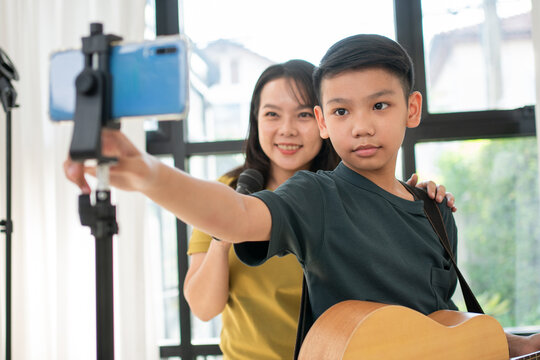 A confident young guitarist records a performance on a smartphone while his mother supports him at home. The lifestyle image represents online education, family encouragement, and creative expression